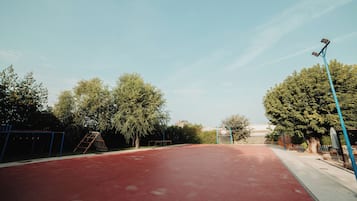 Una cancha de baloncesto al aire libre con superficie roja, rodeada de árboles y una cerca azul.