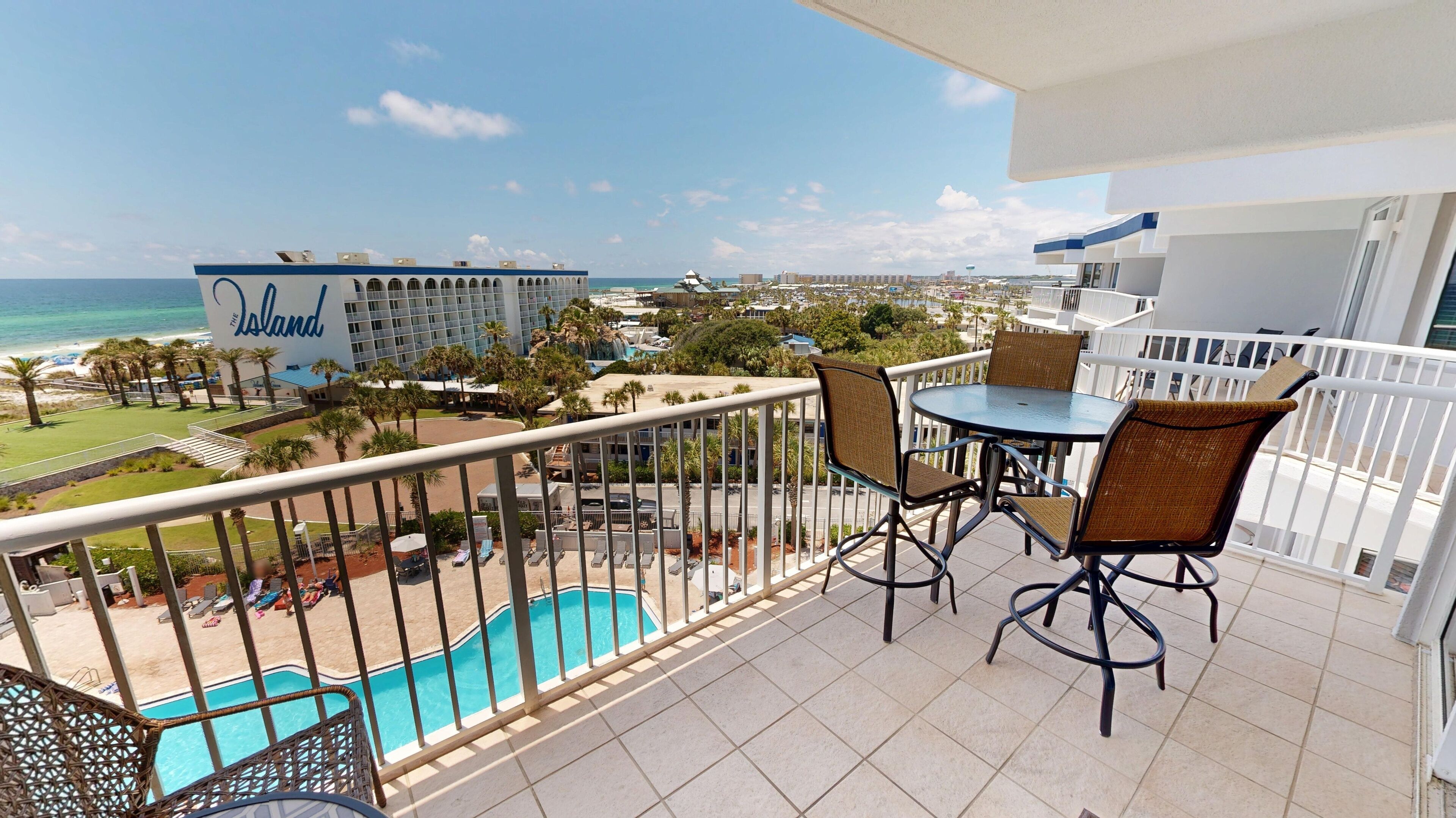 A balcony with a view of a swimming pool, palm trees, and a hotel named "The Island" in the background.