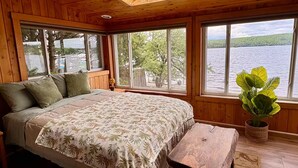 Un dormitorio de cabaña de madera con cama, banco y ventanas amplias con vistas a un lago.