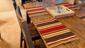 Un comedor con una larga mesa de madera, mantel a rayas y una bandeja decorativa. Hay dos ventanas con cortinas, una silla de madera y un letrero en la pared.
