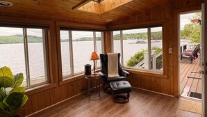 Una habitación de cabaña acogedora con ventanas amplias que dan a un lago, un sillón cómodo, una mesita con lámpara y una planta en maceta.