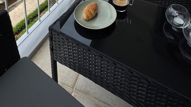 Un balcon avec une table en osier, une assiette avec un croissant et un verre de café. On aperçoit des palmiers et une vue sur un immeuble doté de balcons.