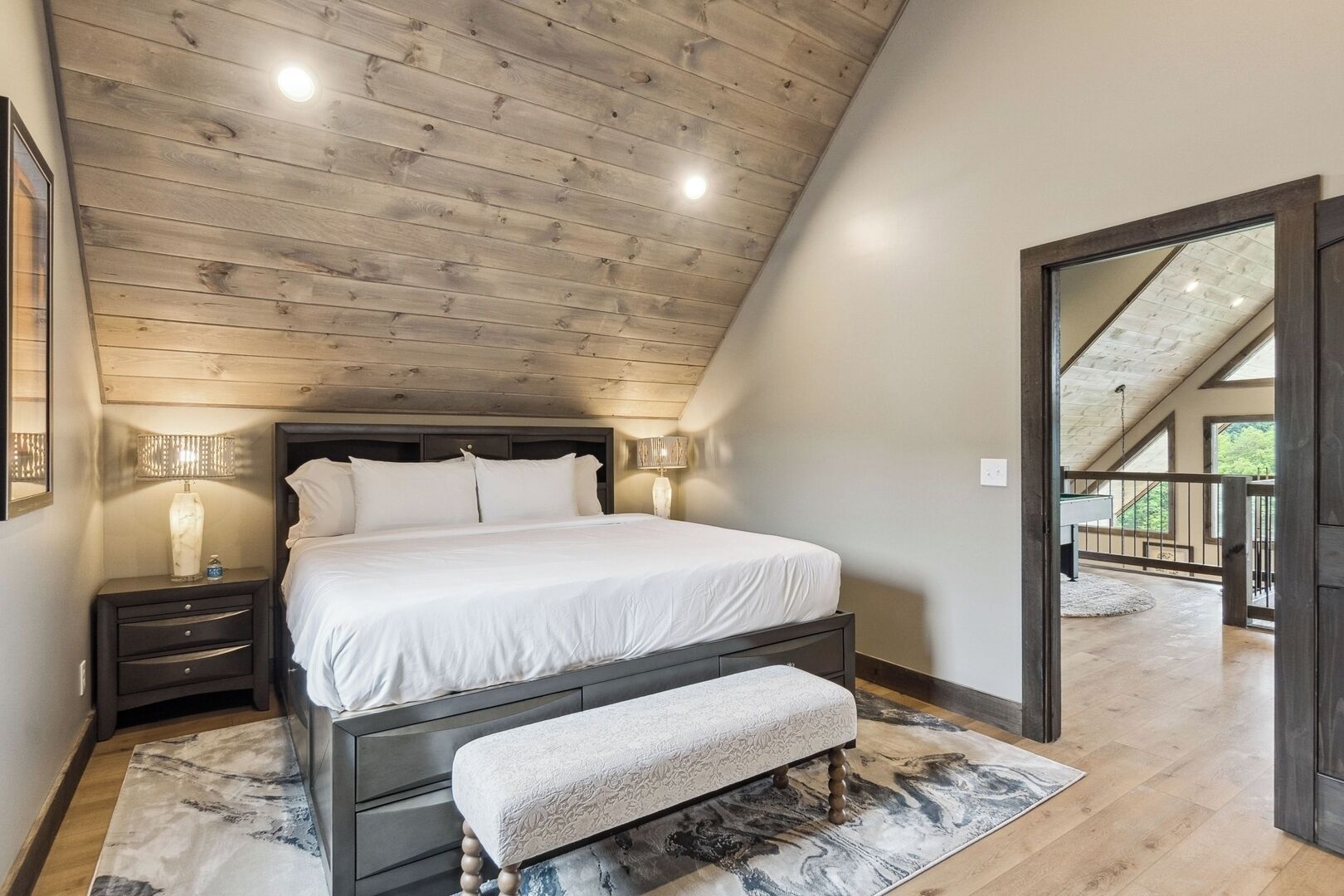Un dormitorio con techo de madera, una cama grande con ropa blanca, lámparas de noche, un banco y vistas a un balcón exterior.