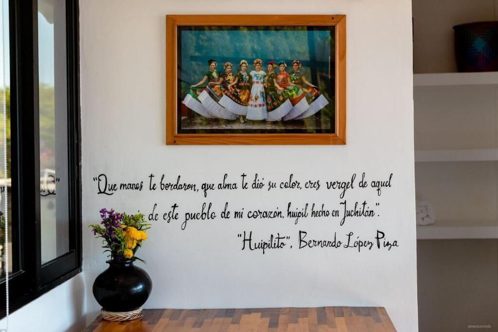 A framed painting of dancers on a wall, a quote in Spanish, and a vase with flowers on a table.