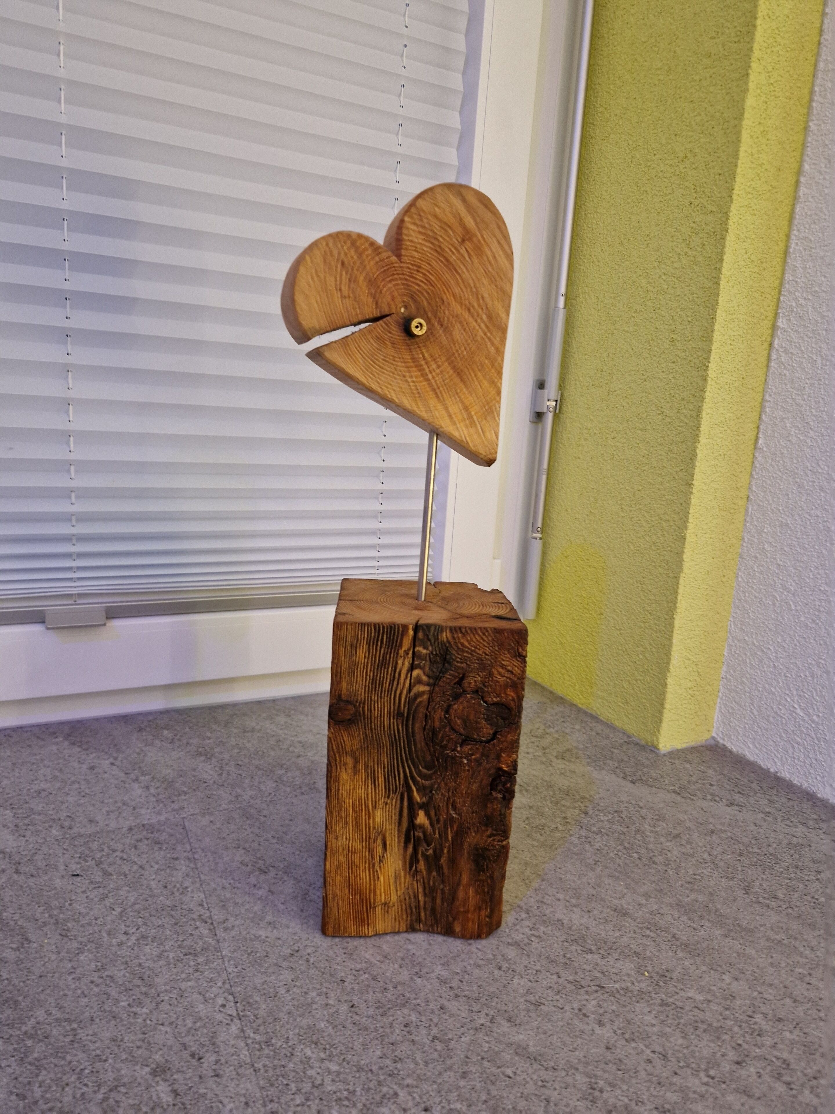 A wooden table with a heart-shaped top and a metal stand, placed on a carpeted floor in a room with a window and a door.