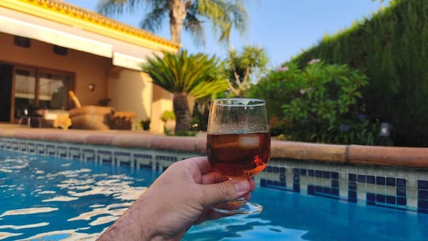 Una mano sostiene un vaso con líquido ámbar sobre una piscina con borde de azulejos y un edificio de techo amarillo al fondo.