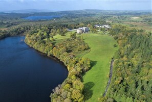 An aerial view of a property with a large body of water, a golf course, and a castle-like building surrounded by dense forests.