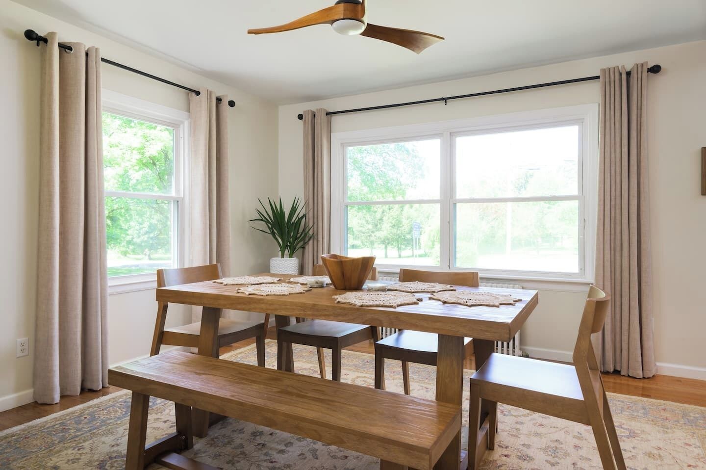 A dining area with a wooden table and benches, large windows with curtains, a ceiling fan, and a potted plant.