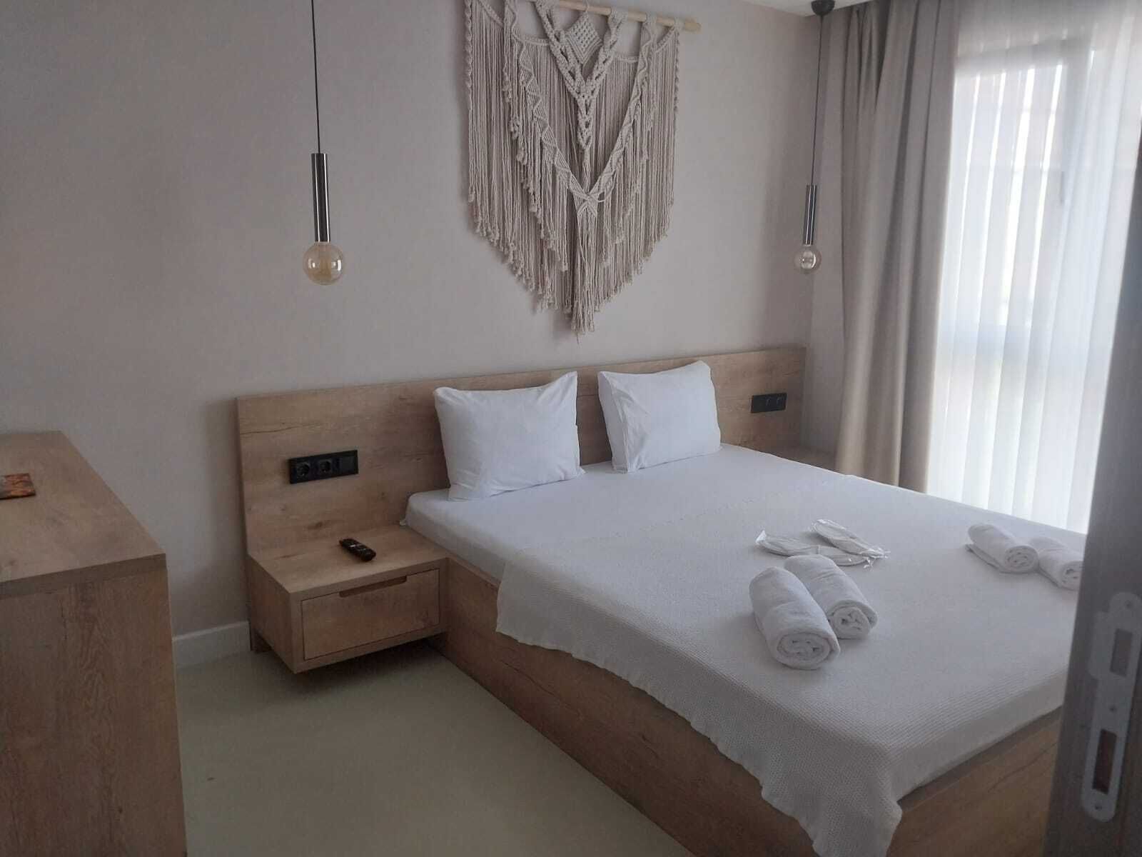 A hotel room with a wooden headboard, a bed with white linens, a macrame wall hanging, and a bedside table with a remote control.