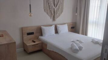 A hotel room with a wooden headboard, a bed with white linens, a macrame wall hanging, and a bedside table with a remote control.