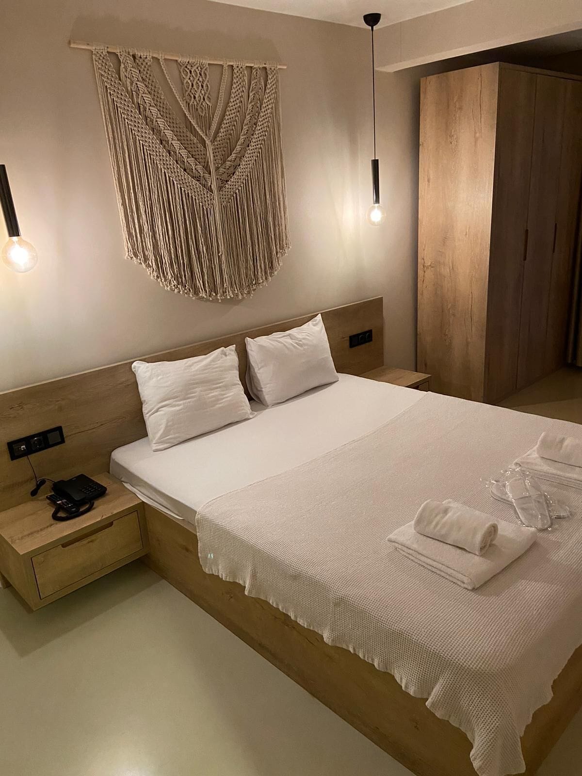 Una cama bien hecha con sábanas blancas, cabecera de madera y un colgador de macramé en la pared. Hay una mesita de noche con un teléfono y una lámpara.