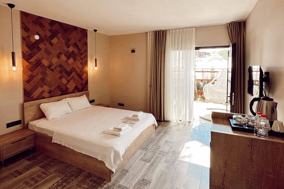 A hotel room with a large bed, wooden headboard, two bedside tables, a nightstand, a desk with a coffee maker, and a window with curtains.