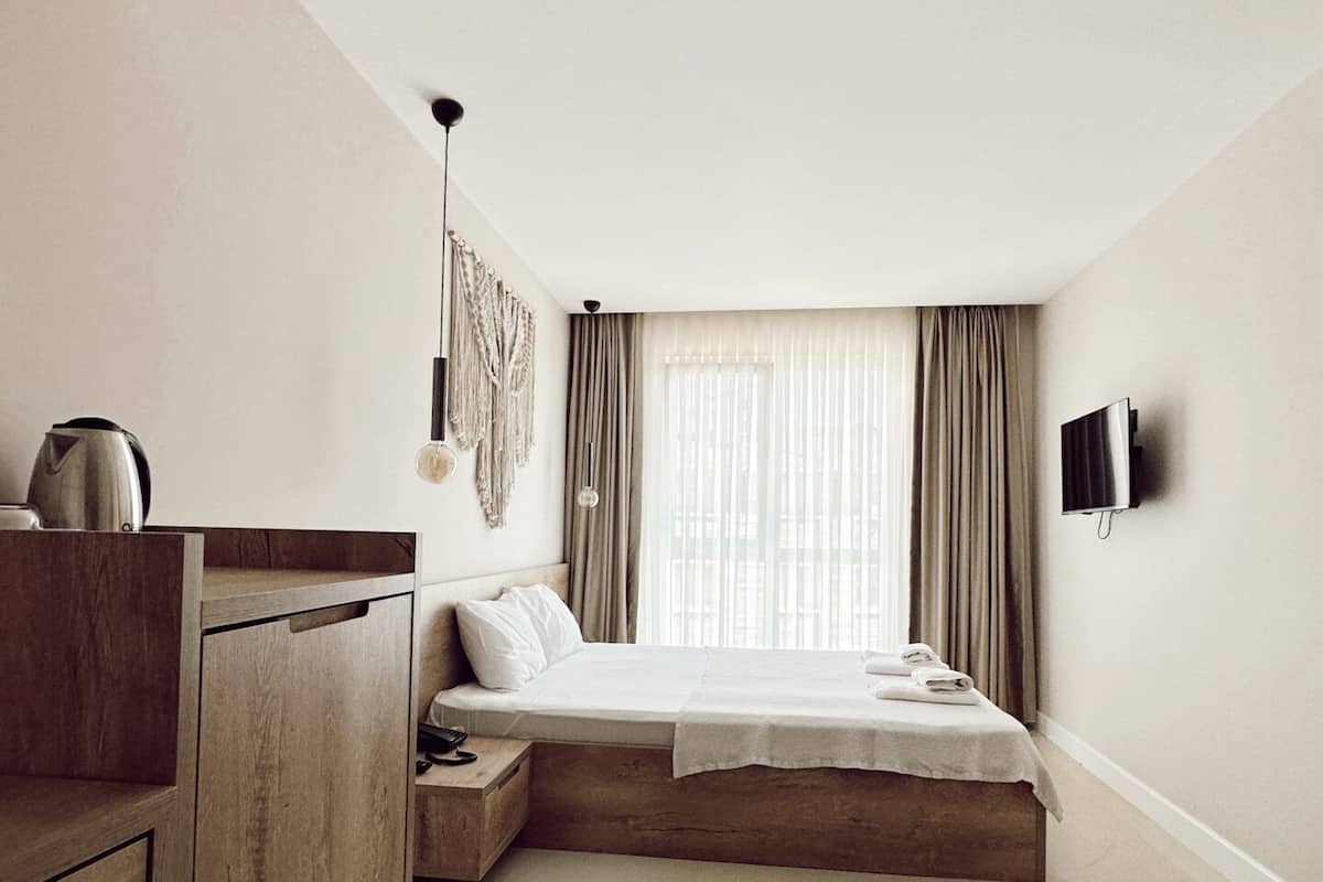 A minimalist bedroom with a bed, a wooden nightstand, a kettle, and a TV mounted on the wall.