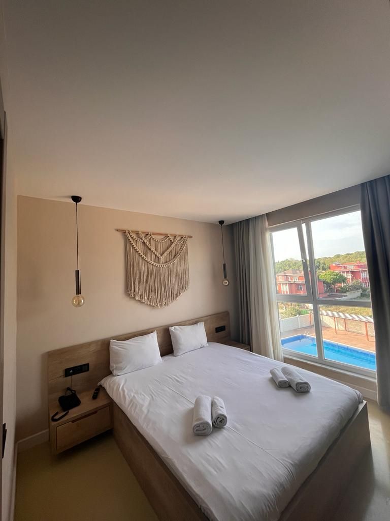 A hotel room with a large bed, a macrame wall hanging, a view of a swimming pool, and a balcony with outdoor seating.