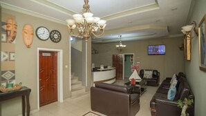 A spacious living room with a brown leather sofa, a coffee table, and a chandelier. There is a reception desk in the background, and a television mounted on the wall. - A Charming Retreat in Lubumbashi (Lubumbashi)