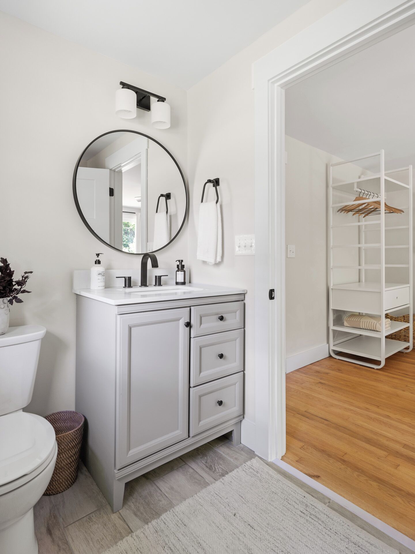 Une salle de bain avec un meuble-lavabo blanc, un miroir rond, un porte-serviettes et un dressing avec des étagères.