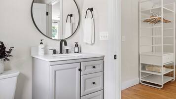 Une salle de bain avec un meuble-lavabo blanc, un miroir rond, un porte-serviettes et un dressing avec des étagères.
