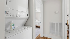 A laundry area with a front-loading washer and dryer, a bottle of Seventh Generation laundry detergent, and a vented air conditioning unit. - Luxury King Suite, 125-yr historic home, 2 blocks from Main St. Crib included (Clayton)
