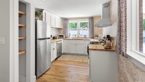 A modern kitchen with white cabinets, a stainless steel refrigerator, a microwave, and a sink. There is a window with patterned curtains and a view of greenery outside. - Luxury King Suite, 125-yr historic home, 2 blocks from Main St. Crib included (Clayton)