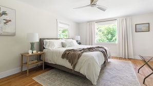 A bedroom with a bed, bedside tables, a ceiling fan, a window with curtains, and a view of greenery outside. - Luxury King Suite, 125-yr historic home, 2 blocks from Main St. Crib included (Clayton)