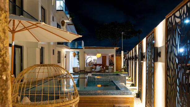 A nighttime view of a modern outdoor pool area with a glass-enclosed hot tub, wicker seating, and a wooden deck. - Ocean Park Beach Club Full (San Juan)