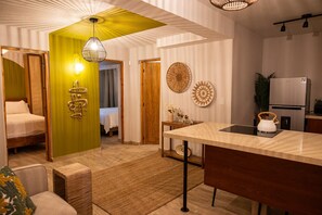 Een moderne hotelkamer met een bed, een zithoek met een bank en een salontafel, een keuken met een koelkast en waterkoker, en een eethoek met een tafel en stoelen.