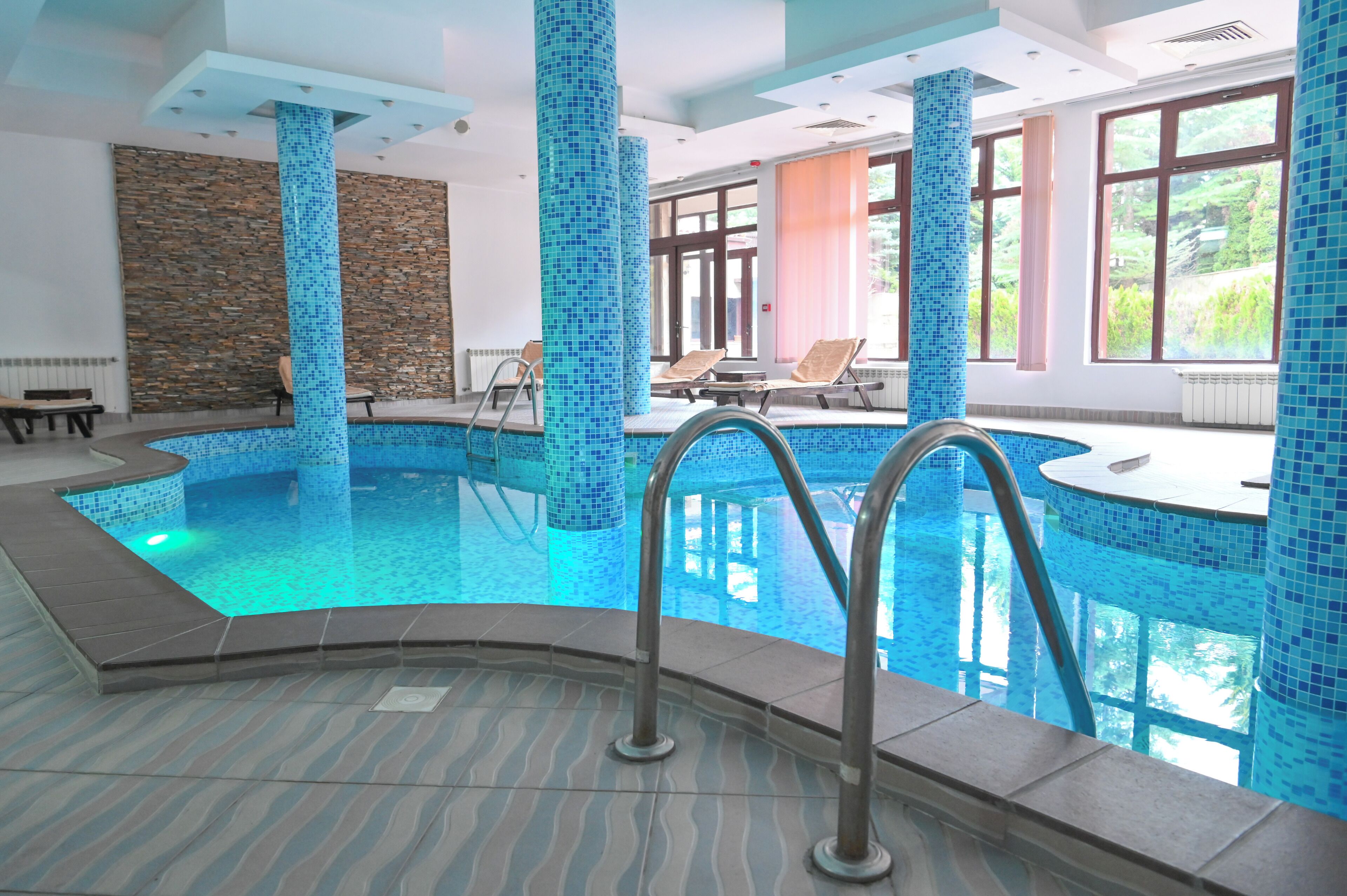 Una piscina interior con azulejos azules de mosaico, sillas de descanso y ventanas amplias.