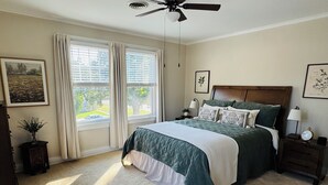 Un dormitorio con cama, dos ventanas con cortinas, ventilador de techo, mesita de noche con lámpara y un cuadro en la pared.