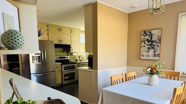 Una cocina moderna con electrodomésticos de acero inoxidable, un comedor con mesa y sillas blancas, y un cuadro en la pared.