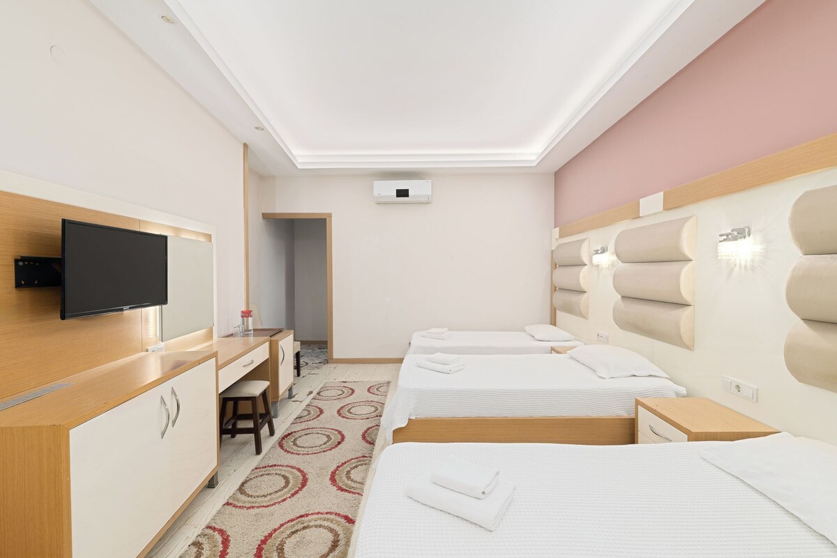 A hotel room with two beds, a desk with a flat-screen TV, a small seating area, and a bathroom visible through an open door.
