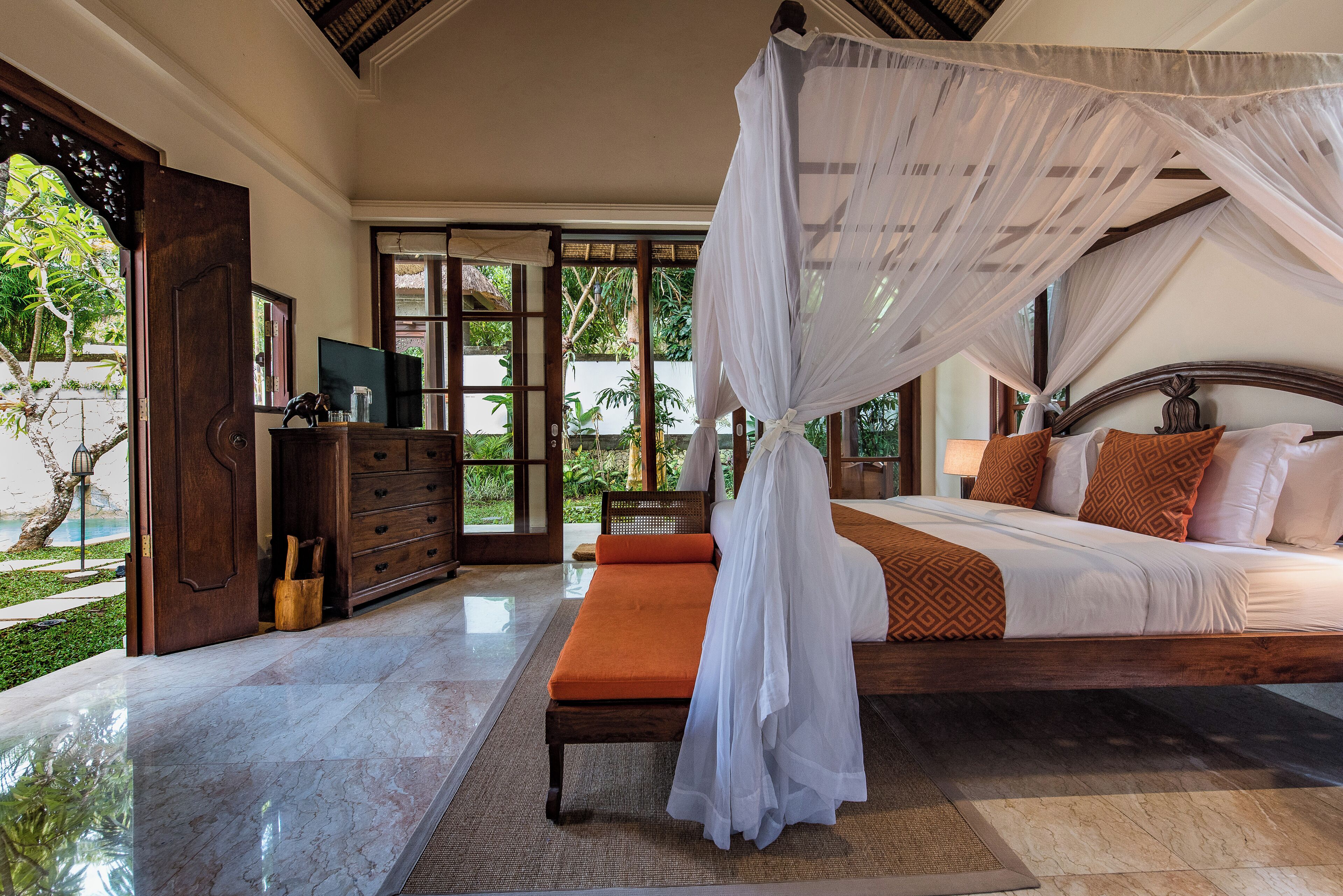 A bedroom with a canopy bed, a wooden bench, and a view of the garden through the door.