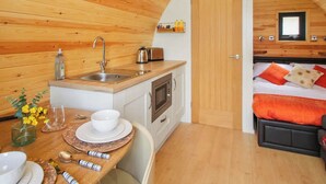 A compact room with a wooden ceiling, a kitchenette with a sink and stove, a bed with orange bedding, and a small dining area with a table set for two.