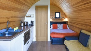 A compact room with a wooden ceiling, a kitchenette, a bed with colorful bedding, and a small window.