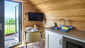 A wooden interior with a dining table, chairs, a TV mounted on the wall, a sink, and a window overlooking a green landscape.