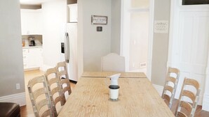Comedor con mesa de madera, sillas blancas, ventilador de techo y cocina al fondo.