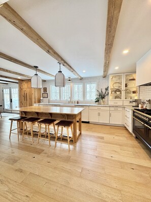 A spacious kitchen with wooden beams, a central island with stools, white cabinets, and a large window. - Tucked-Away 3 Bedroom Farmhouse Cottage 3 Private Acres (Arundel)