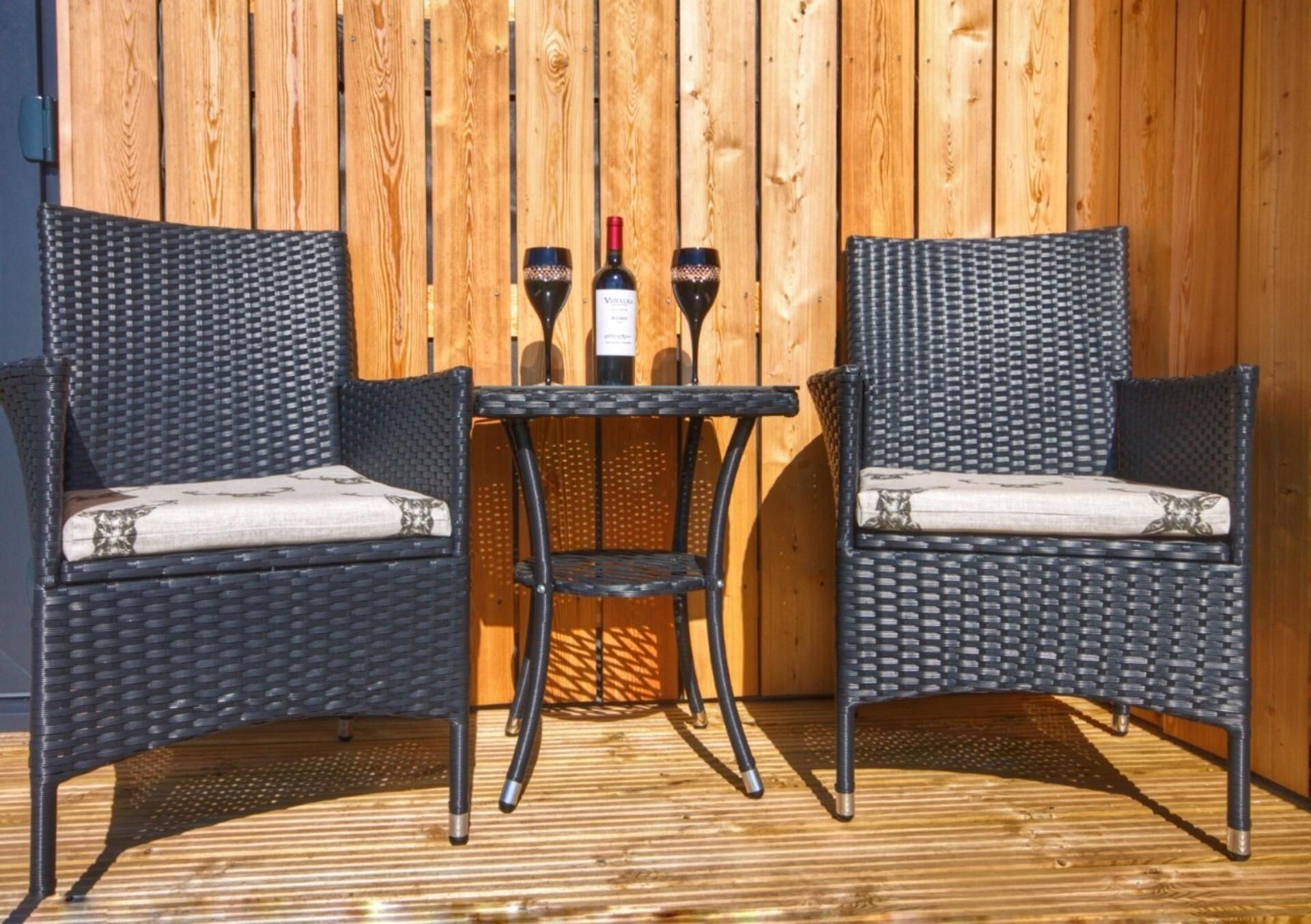 Two wicker chairs with cushioned seats and backs, a small table, and two wine glasses with bottles on a wooden deck.