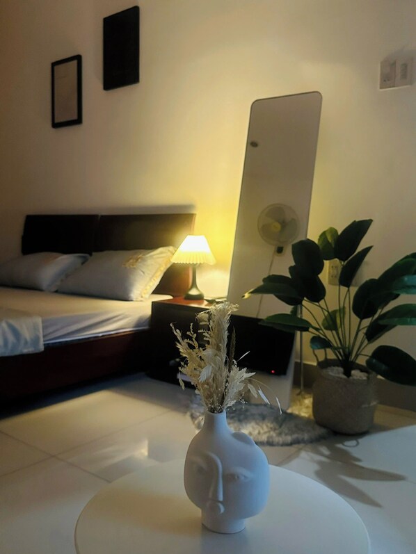 Una habitación de hotel con cama, mesita de noche con lámpara, un espejo grande y dos plantas en macetas.