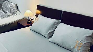 A neatly made bed with a white sheet and two pillows, one with a gold emblem. There is a bedside table with a lamp and a power outlet. The wall has two framed pictures, one of a coffee cup and another of a mug.