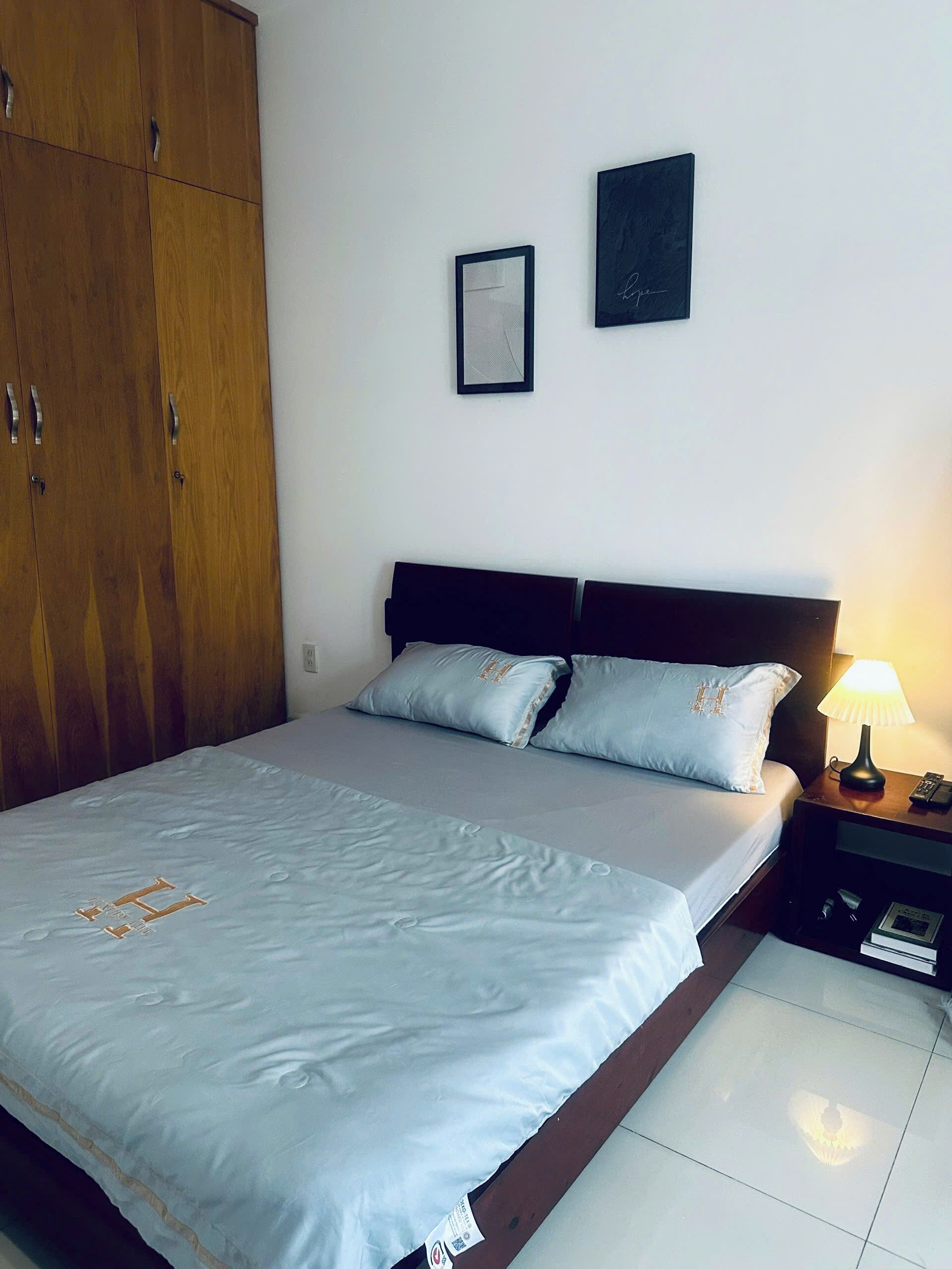 A hotel room with a bed, a nightstand with a lamp, a framed certificate on the wall, and a wooden wardrobe.