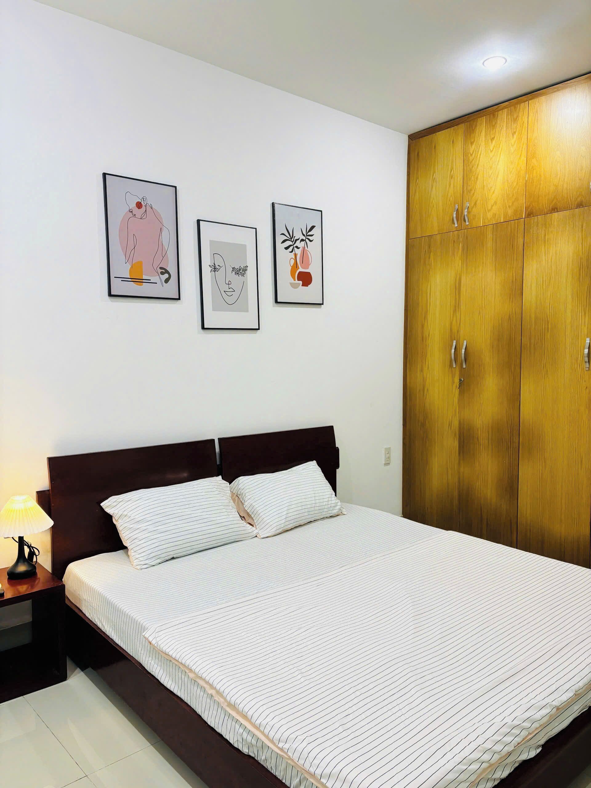 Una cama bien hecha con ropa de cama a rayas, un armario de madera y tres cuadros en la pared.