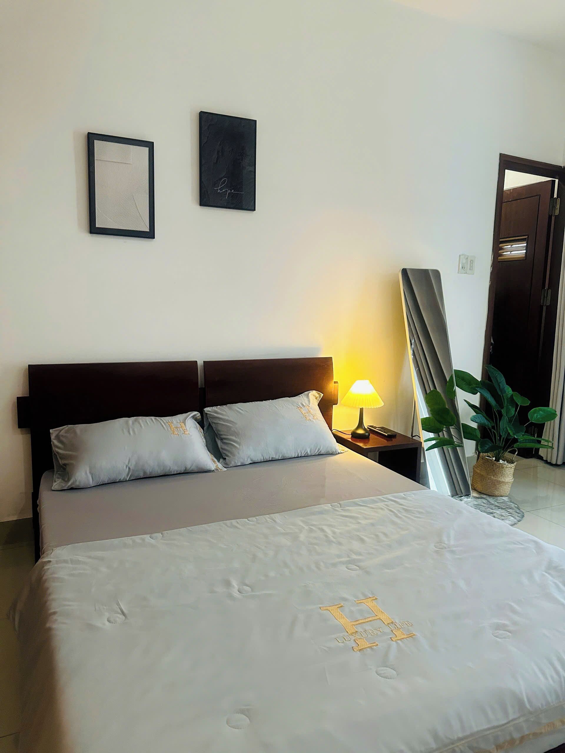 Hotelkamer met een bed, twee kussens, een nachtkastje met een lamp, een lijst en een potplant.
