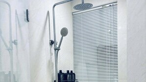 A modern bathroom with a glass-enclosed shower, a handheld showerhead, a wall-mounted faucet, and a window with blinds. - The Cozy Home (Nha Trang)