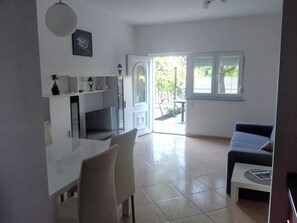 Una sala moderna con sofá, una mesa de comedor y una televisión. Hay una zona de cocina con armarios y también una televisión. La habitación tiene ventanas amplias y vistas al exterior.