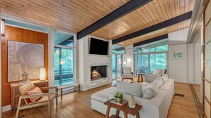 A modern living room with a fireplace, wooden flooring, and large windows offering a view of the outdoors.
