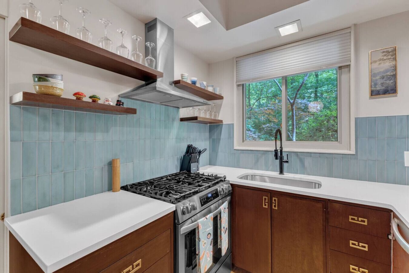 A kitchen with a gas stove, oven, and a window with a view of trees. There are wooden cabinets and a white countertop.