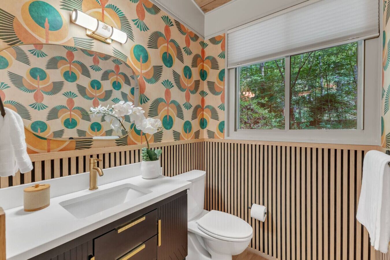 A modern bathroom with a wooden slat wall, a white sink with gold handles, a toilet, and a window with a view of trees.