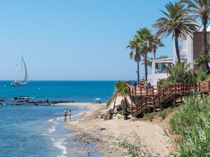 A beach with a wooden walkway leading to a building, palm trees, and a sailboat in the distance.