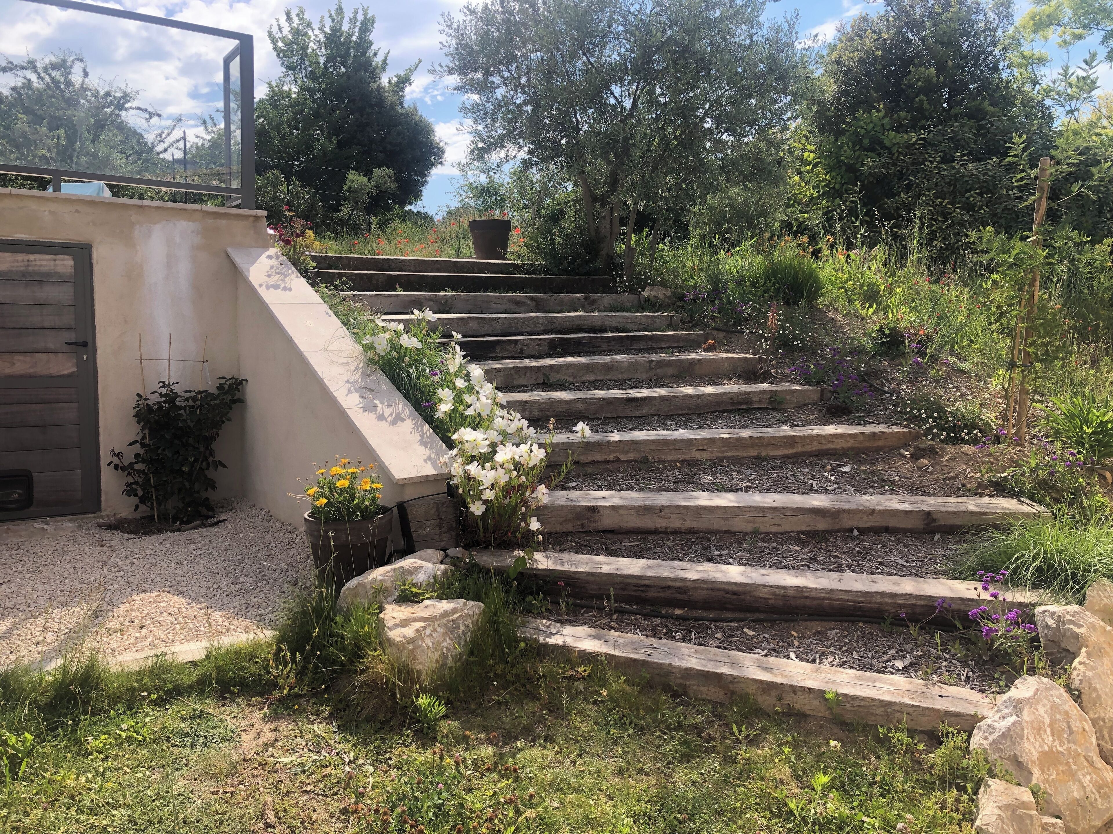 Eine Steintreppe führt zu einem Gebäude mit einer Tür, umgeben von einem Garten mit verschiedenen Pflanzen und Blumen.