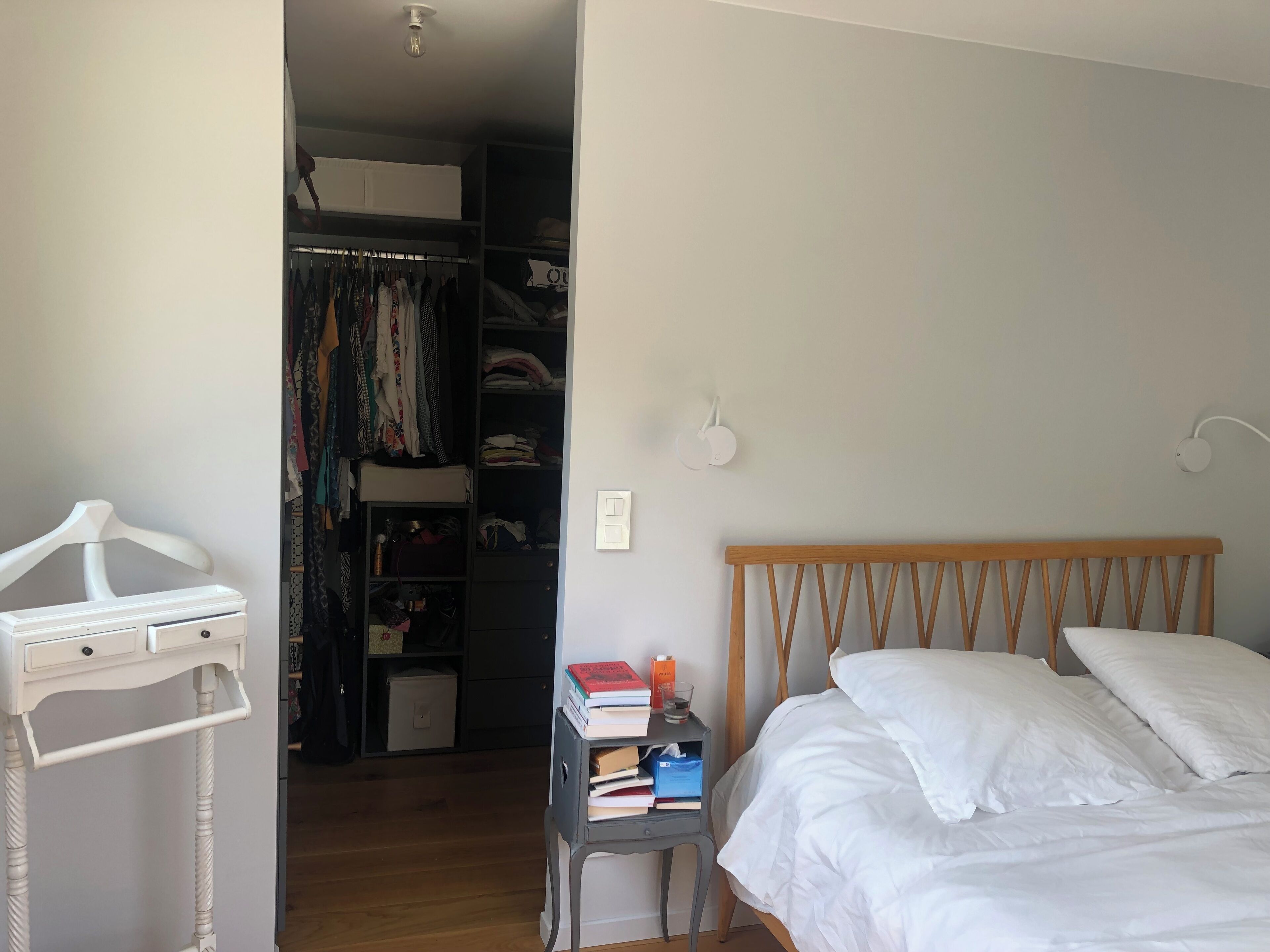 A bedroom with a wooden bed, white pillows, and a bedside table with books and a glass. A white dresser with drawers is on the left, and a closet with hanging clothes is in the background.